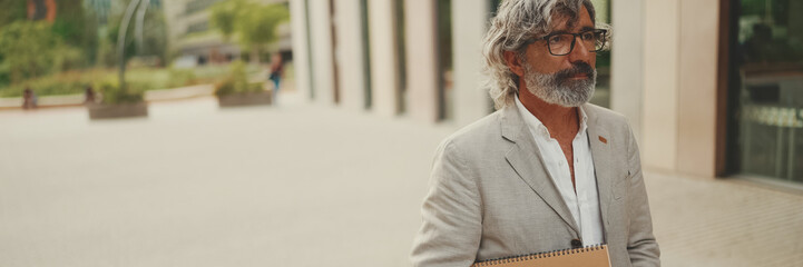 Panorama of  mature businessman with beard in eyeglasses wearing gray jacket looks around and walks down the street past modern buildings