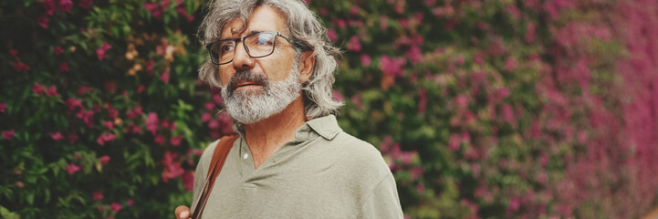 Friendly middle-aged man with gray hair and beard, walks next to wall with purple flowers blooming in the background, Panorama