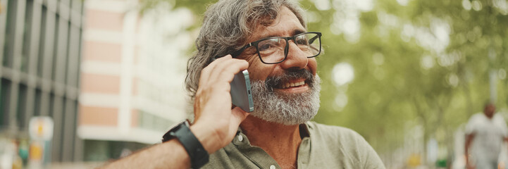 Laughing middle-aged man with gray hair and beard wearing casual clothes sits on bench and uses...