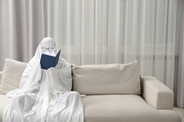 Creepy ghost. Person covered with white sheet reading book on sofa at home, space for text