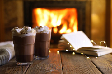 Glasses with hot cocoa, marshmallows, lights and book on wooden table near fireplace