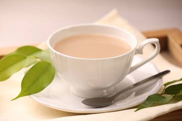 Cup of aromatic tea with milk, spoon, saucer and green leaves on table, closeup