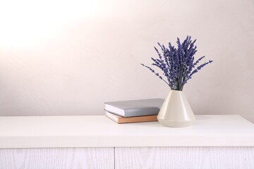 Bouquet of beautiful preserved lavender flowers and notebooks on white wooden chest of drawers near beige wall, space for text