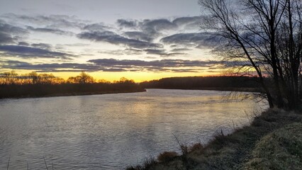 On a winter morning, the sun has not yet risen above the trees on the far bank of the river around...