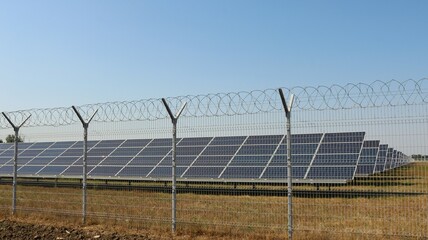 a complex of solar panels behind a barbed wire fence in a field under a clear blue sky, a country solar power station behind a guarded barrier, solar energy in industry