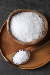 Natural salt in wooden bowl and spoon on dark grey table, top view