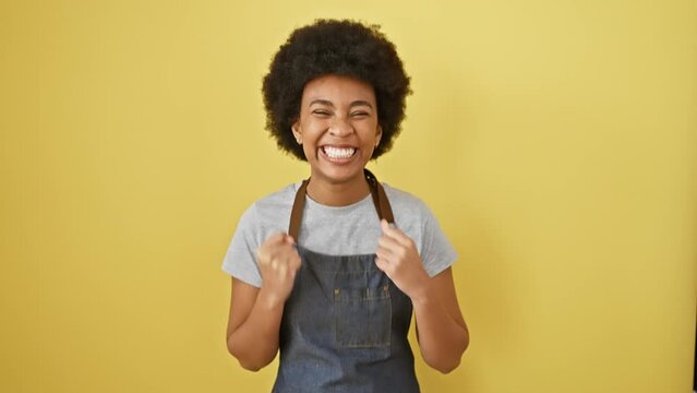 Joyful african american woman in apron celebrates victory, screaming with excitement and happiness while making winner gesture on yellow isolated background