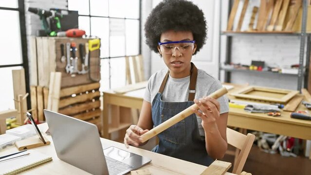 A focused woman inspects wood while multitasking with a laptop in a carpentry workshop.