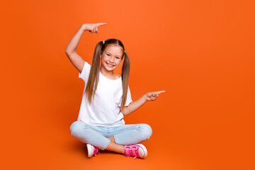 Full length photo of positive nice schoolchild sit floor indicate fingers empty space isolated on orange color background