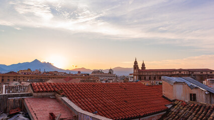 View of the mountains above the rooftops against the setting sun and pink cloudy sky. Red clay...