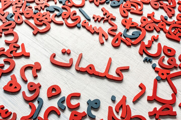 Topview picture of a group of Arabic letters made of wood, with the word (klmat) in the middle, colored red and blue on a gray background.