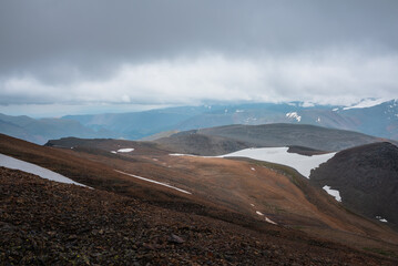 Dark overcast landscape with high pass, wide glacier on stony hill slope and snowy mountain range silhouette in far away in rainy low clouds. Large mountains with snow under gray sky in bad weather.