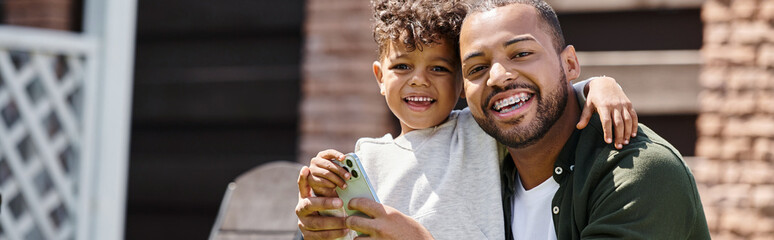 positive african american man in braces and son using smartphone on backyard of house, banner