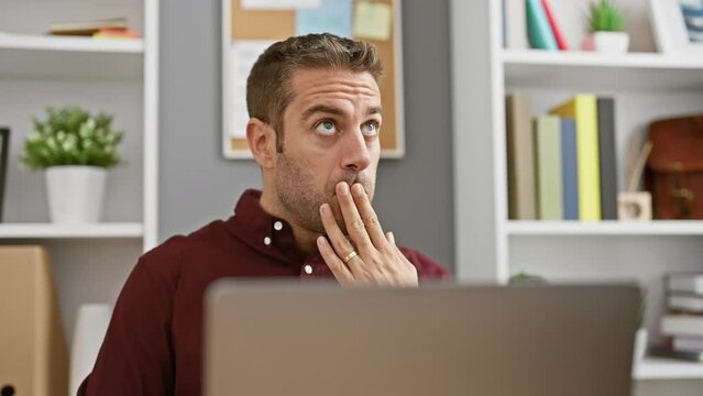 Surprised young hispanic man, scared in silence at the office, covers mouth with hand in shock while sitting at computer
