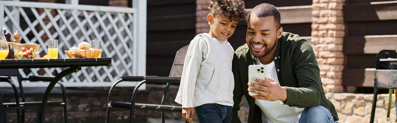 happy african american father and son looking at smartphone and standing on backyard, banner - 740861476