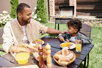 cheerful african american father eating lunch with curly son outdoors, grilled sausages and corn