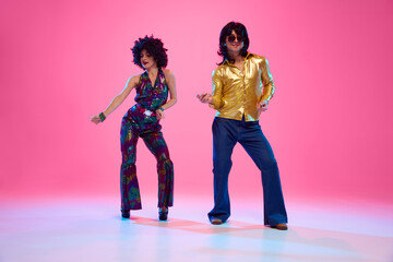 Man and woman dressed in vibrant 1970s attire dancing in motion against gradient pink studio...