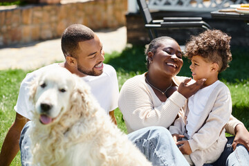 family portrait of cheerful african american family smiling and sitting on lawn near dog