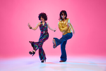 Retro disco era fashion. Couple with funky hairstyles and groovy clothes dancing energetic dance against gradient pink studio background. Concept of American culture, 1970s, 1980s fashion, music.