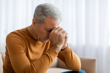 Sad elderly man sitting on sofa at home, touching head