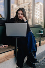 Portrait of cute Korean girl in cafe near window and networking, looking at laptop and drinking coffee. Student having cappuccino and working on laptops outdoors