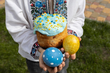 two painted yellow-blue eggs and Easter homemade Easter cake in the hands of a Ukrainian child in...