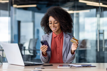 Satisfied african american female keeping debit card and cellular phone in hands by desk with computer and notebook. Pleased lady entering bank data in application for completing cashless payment - obrazy, fototapety, plakaty