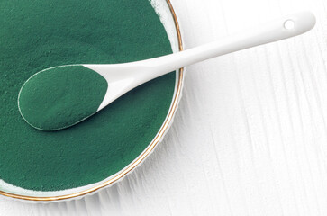 Green chlorella and spirulina powder in a ceramic spoon on a white wooden background, from above view