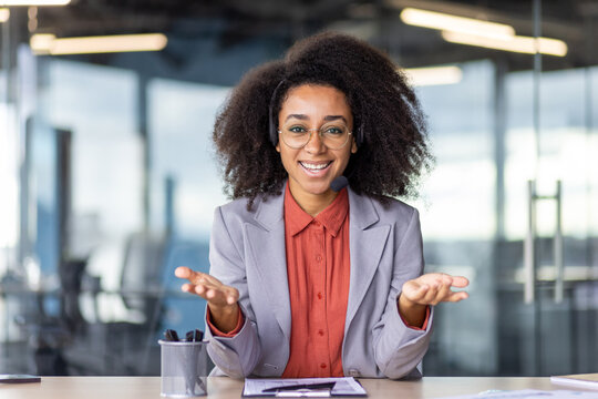 Smiling Latino Woman In Headphones Sitting By Table In Front Of Clipboard With Raised Hands On Blurred Background. Happy Call Centre Operator Gesturing Arms While Hosting Online Video Meeting