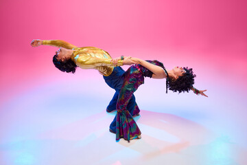 Top view. Couple wearing 70s disco fashion in dramatic back bend dance move against gradient pink studio background. Concept of American culture, 1970s, 1980s fashion, music, comparisons of eras.