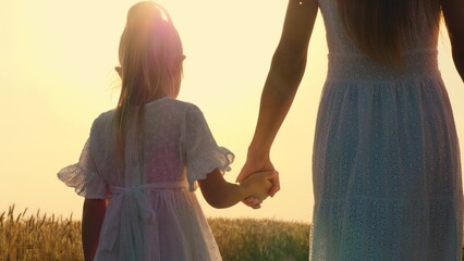 Mother and daughter walking at sunny autumn wheat field holding hands with love tenderness back...