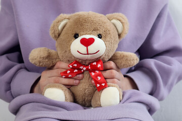 Soft toy - a bear cub with a red ribbon in the hands of a girl on a background of pastel clothes, close-up