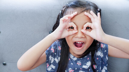 Young Asian girl over gray couch background doing ok gesture like binoculars, eyes looking through...