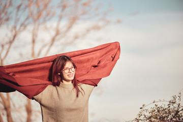 Happy joyful teenage girl stands in park raising her red coat above head with her hands.