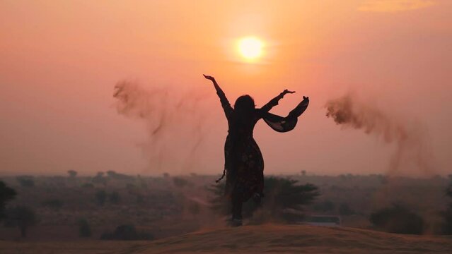 Silhouette shot of Desert sand falling from hands of girl with arm raised against sunset sky in Thar desert at Jaisalmer, Rajasthan, India. Indian girl wearing traditional clothes - Salwar suit