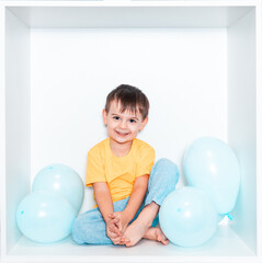 A cute little boy in a green T-shirt and blue pants sits in a white niche of furniture in his room....