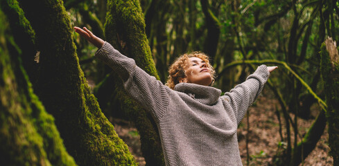 Celebrating freedom and nature love. One woman outstretching arms in the forest with green trees trunk and musk in background. Happiness and wellbeing in natural healthy lifestyle people in leisure - obrazy, fototapety, plakaty