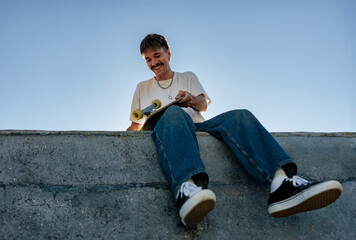 Fototapeta na wymiar Low angle of young male skateboarder holding skateboard sitting on ramp in skate park 