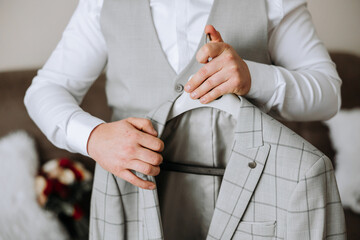 An elegant groom in a white shirt and a stylish tie is holding a jacket on a hanger. Details of the...