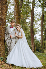 Fototapeta na wymiar lovely and stylish newlyweds are hugging and smiling against the background of autumn nature in a beautiful garden. An incredibly beautiful young bride leaned against the shoulder of her beloved groom