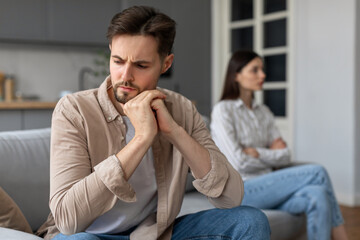 Unhappy young man sitting on sofa separate from discontented wife on background - obrazy, fototapety, plakaty