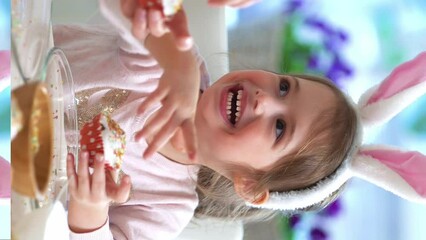 Young mother and her cute little daughter wearing funny rabbit ears are cooking Easter cupcakes sitting at a festive table with basket, eggs and Bunny