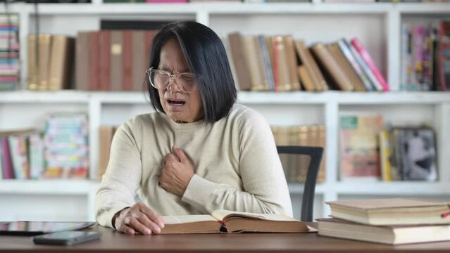 Senior adult elderly woman asian sits in a chair hand holding her chest with disease chest pain from a heart attack in her house. Health care and medical concept
