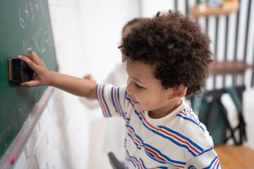 Happy African American kid boy studying and writing with chalkboard and friend in classroom
