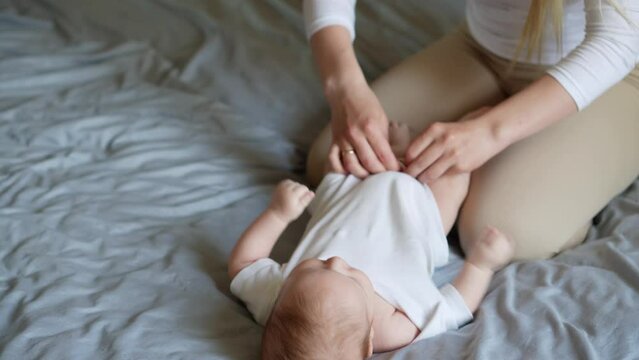 Mother dresses her newborn baby lying on the bed. Woman fastens the buttons on a white bodysuit. Concept of children's hygiene, caring, loving parents.