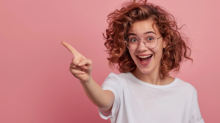 An exuberant young woman with curly hair and glasses, pointing her finger to the side with a wide, joyful smile on a pink background.