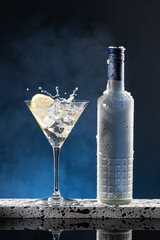 Vodka Cocktail glass splash and ice-cold bottle on blue degraded background with copy space. Vertical format.