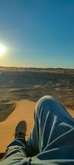 A man's legs stretched out on the sandy crest of a sand dune, watching the sun set on the horizon,...