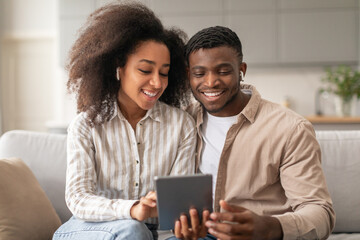 Obraz premium Smiling African couple with wireless earphones using digital tablet indoors