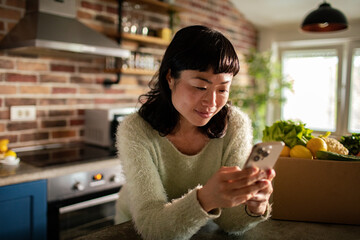 Happy Asian woman using smartphone in kitchen with fresh groceries
